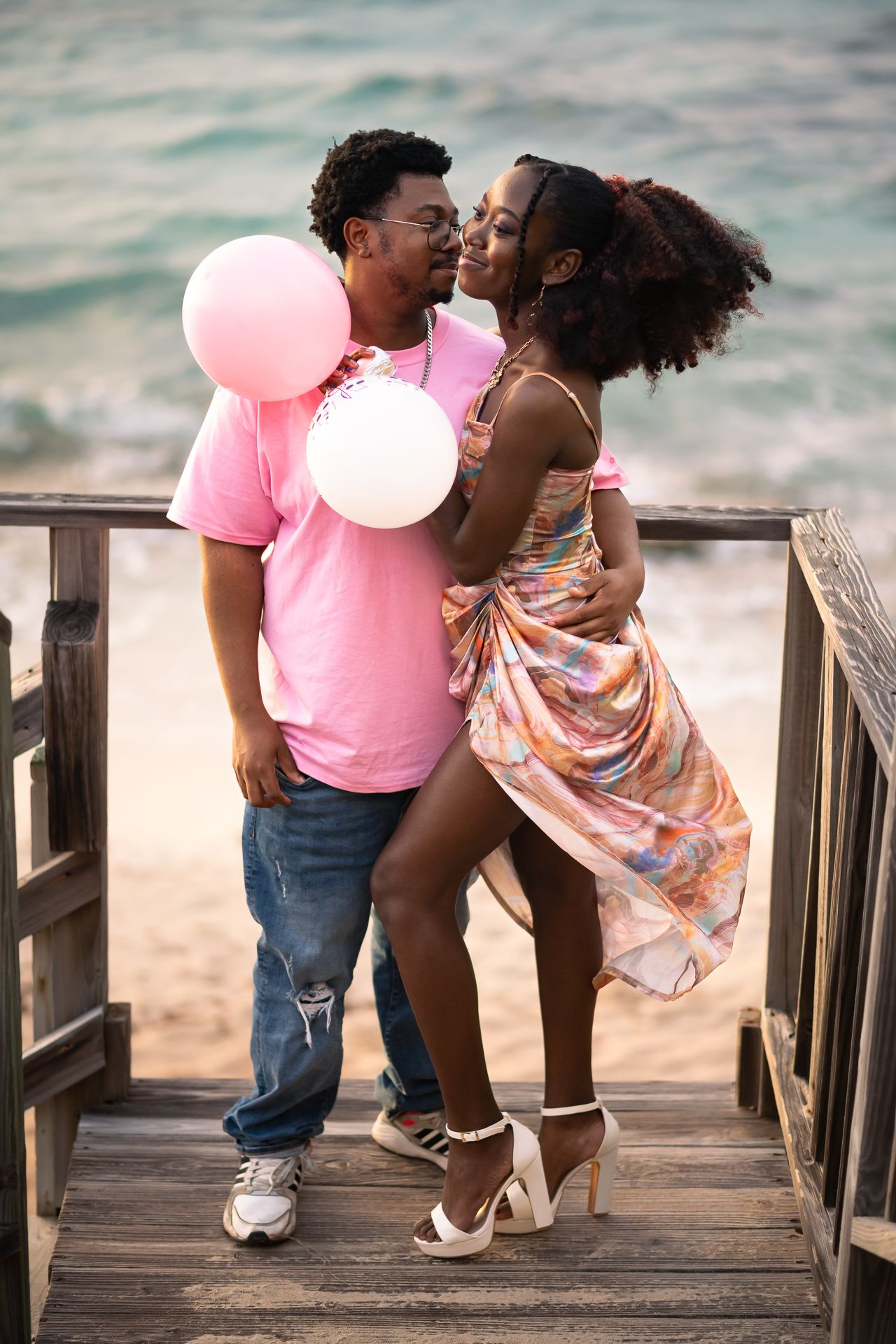 A romantic beach scene with pink balloons and a couple on a wooden boardwalk at sunset.