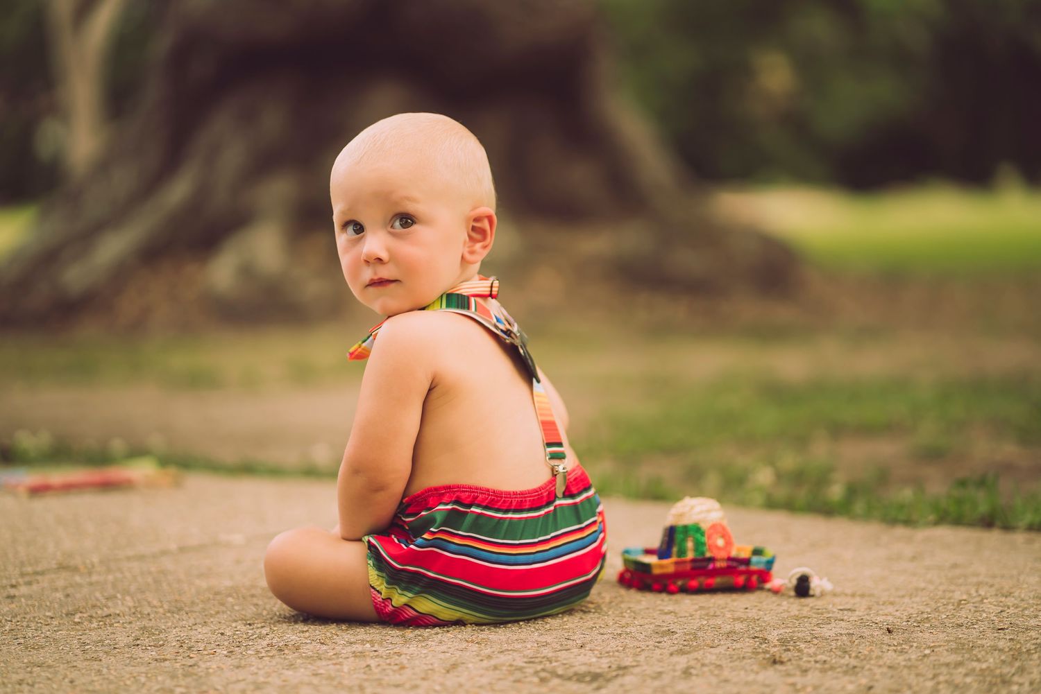 Baby in striped rainbow romper sits on dirt path in outdoor setting.