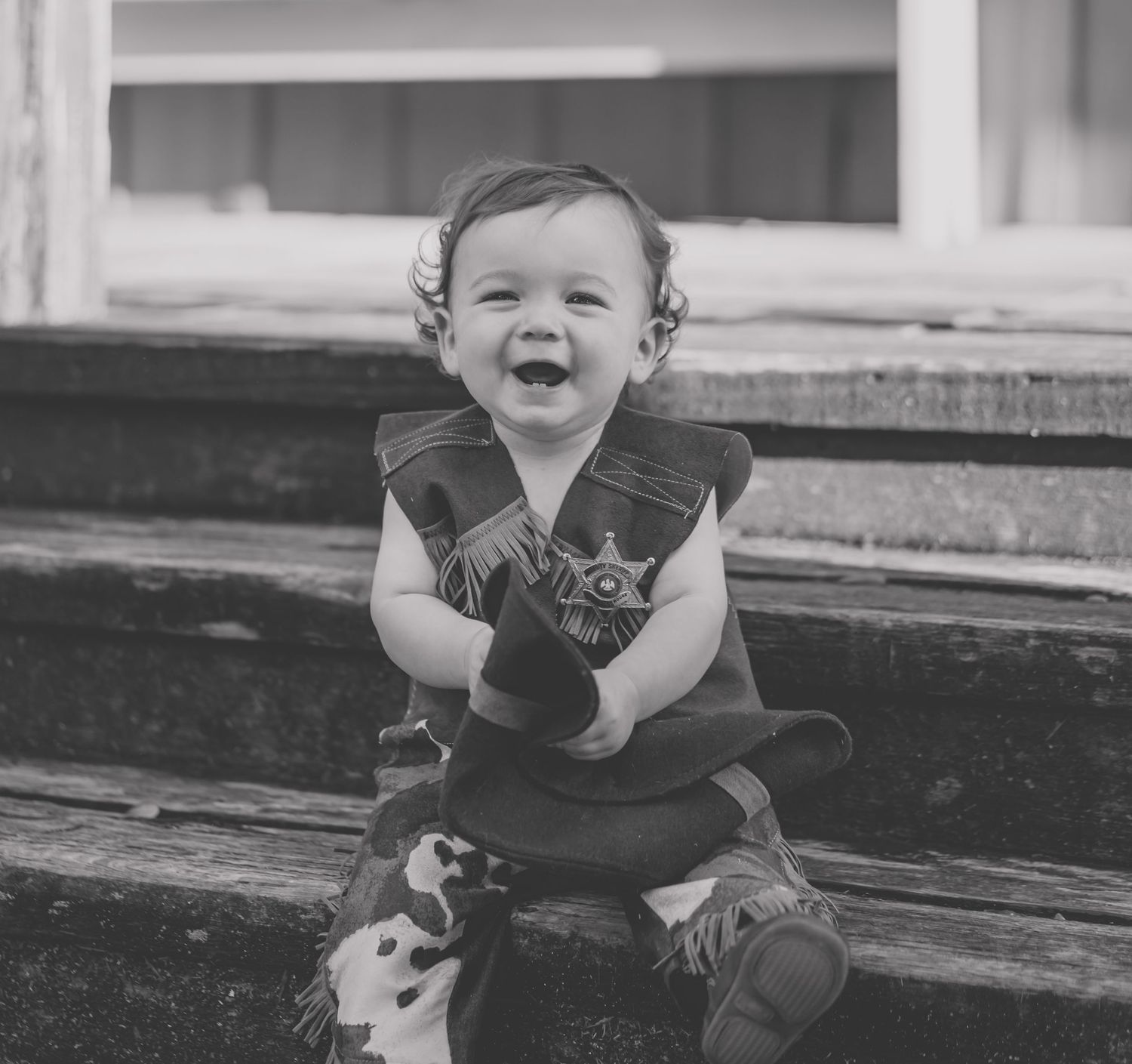 A joyful baby in western wear laughs while sitting on wooden steps in black and white.