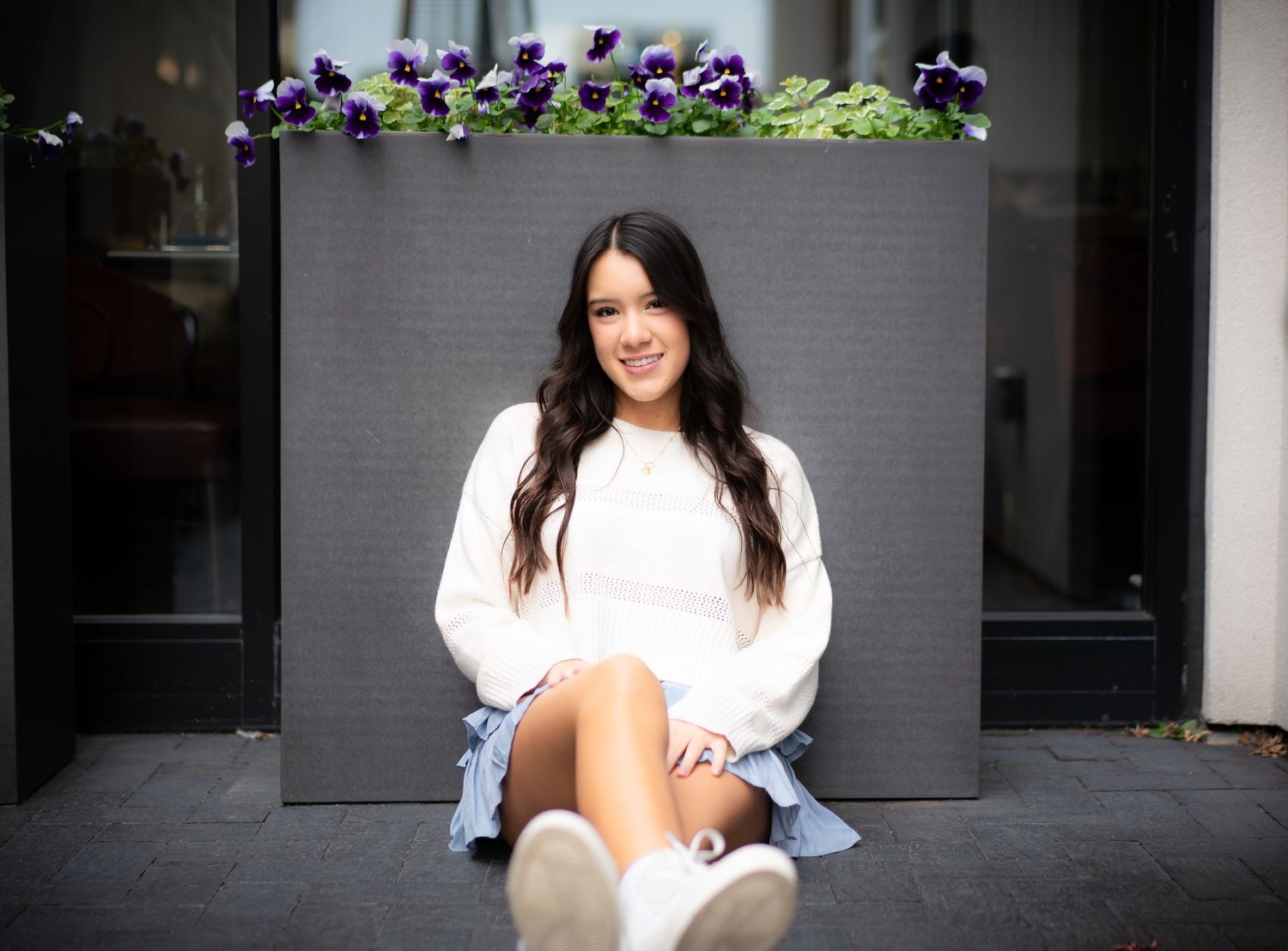 Purple flowers bloom above a concrete wall while a person in white sits on steps below.