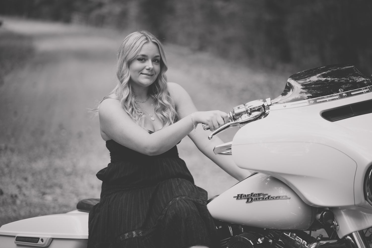 Black and white portrait of someone in a dark dress sitting beside a white motorcycle on a rural road.