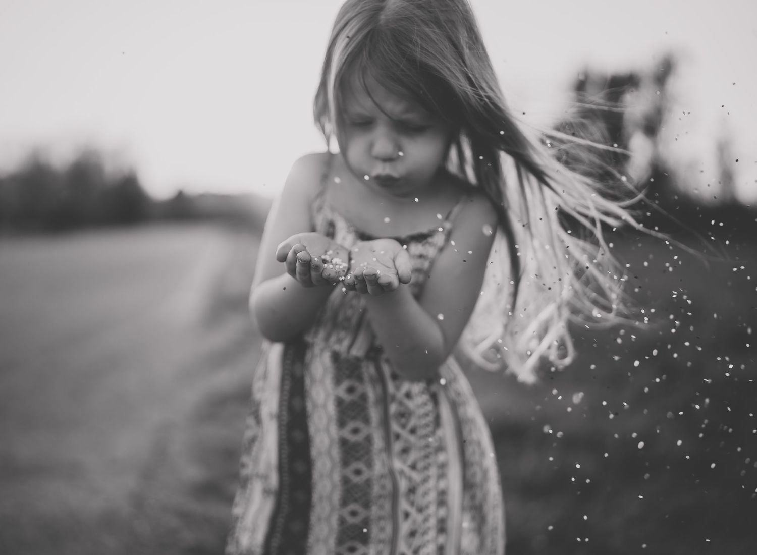 Black and white image of a child blowing dandelion seeds in a summer dress with wind blowing through their hair.