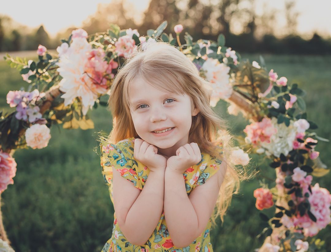 Child poses under floral arch with pink and white flowers in spring garden setting.