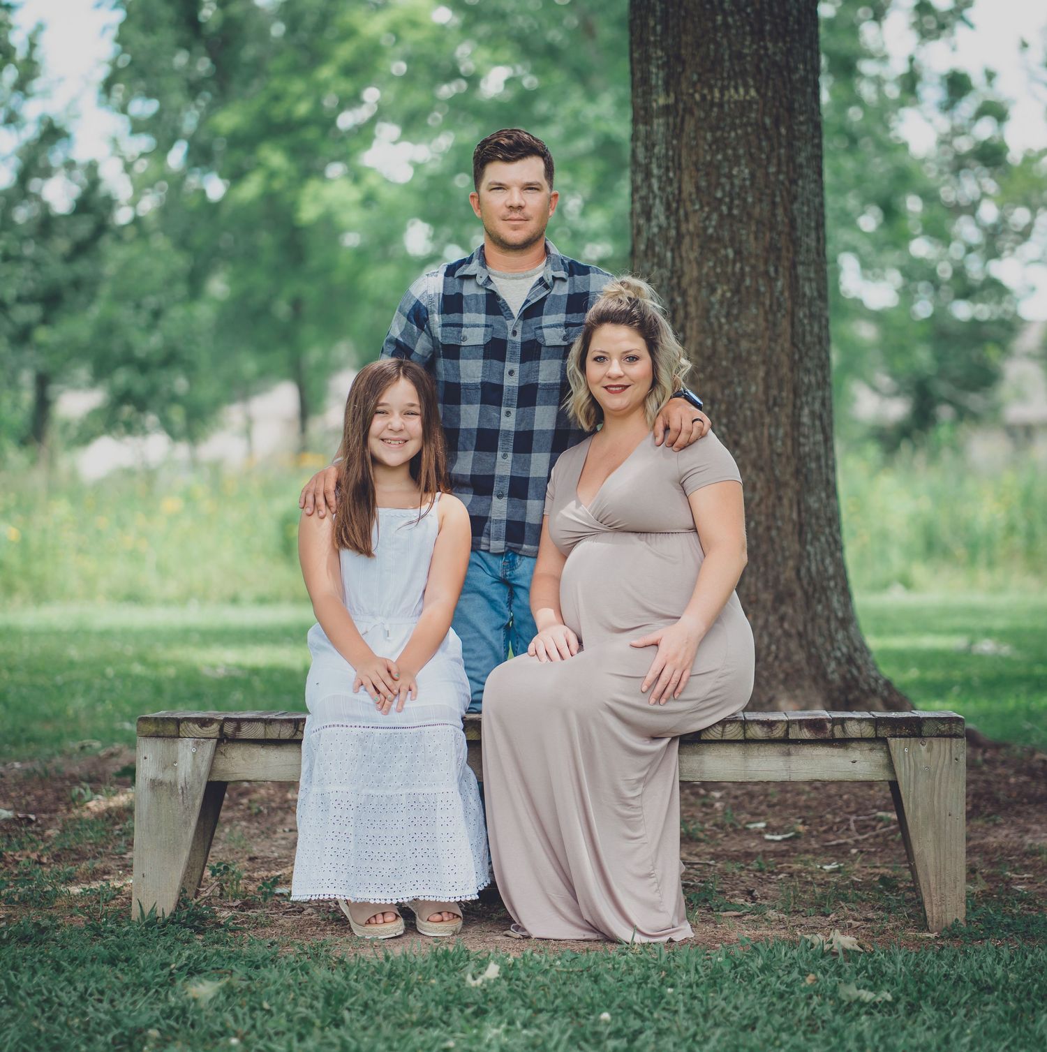 Family sits together on wooden bench under large tree in park setting.