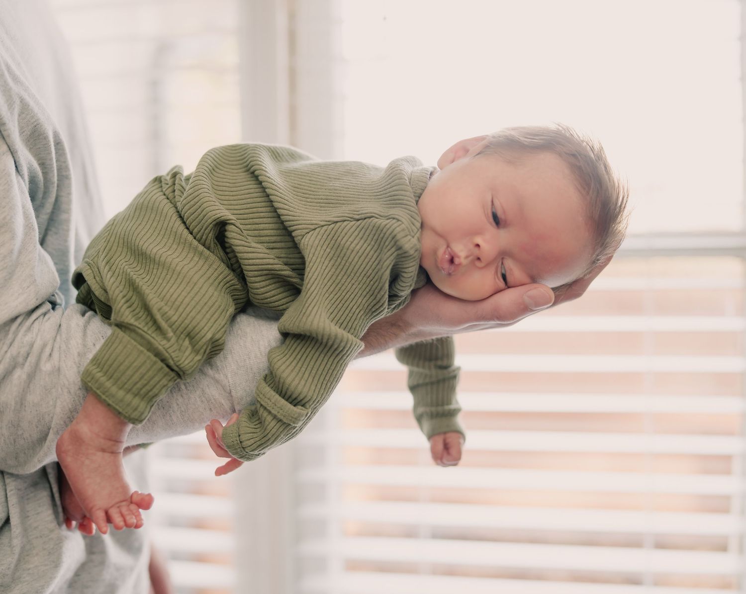 Sleeping baby wearing olive green sweater being held against bright window with horizontal blinds.