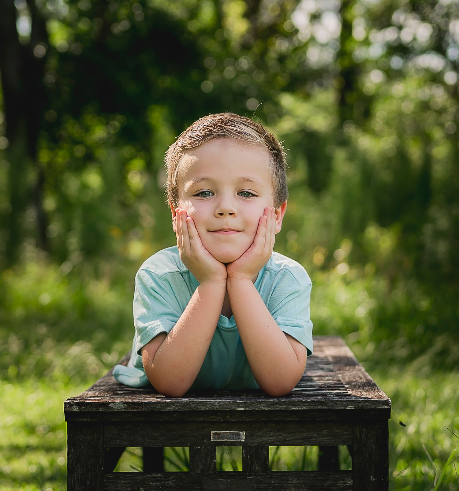 Child in blue shirt resting head on hands while posing on wooden surface outdoors.