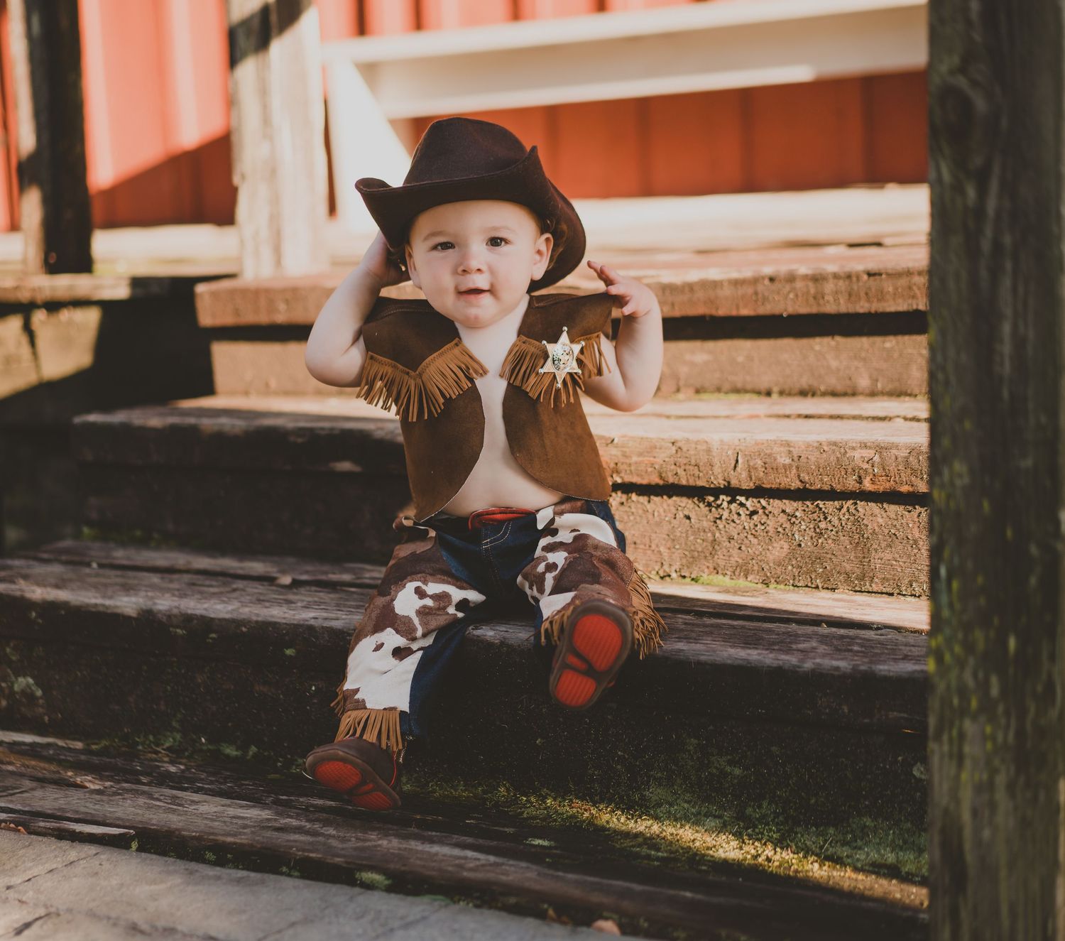 A baby dressed as a cowboy sits on outdoor wooden steps wearing a hat and fringe vest.