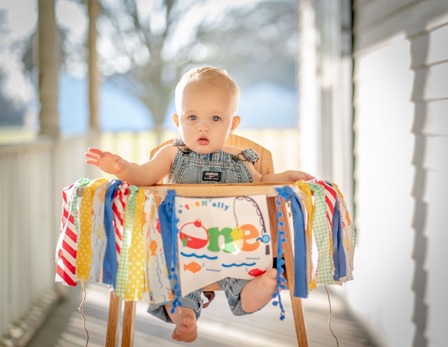 Baby sitting in decorated high chair with colorful ribbons on porch.