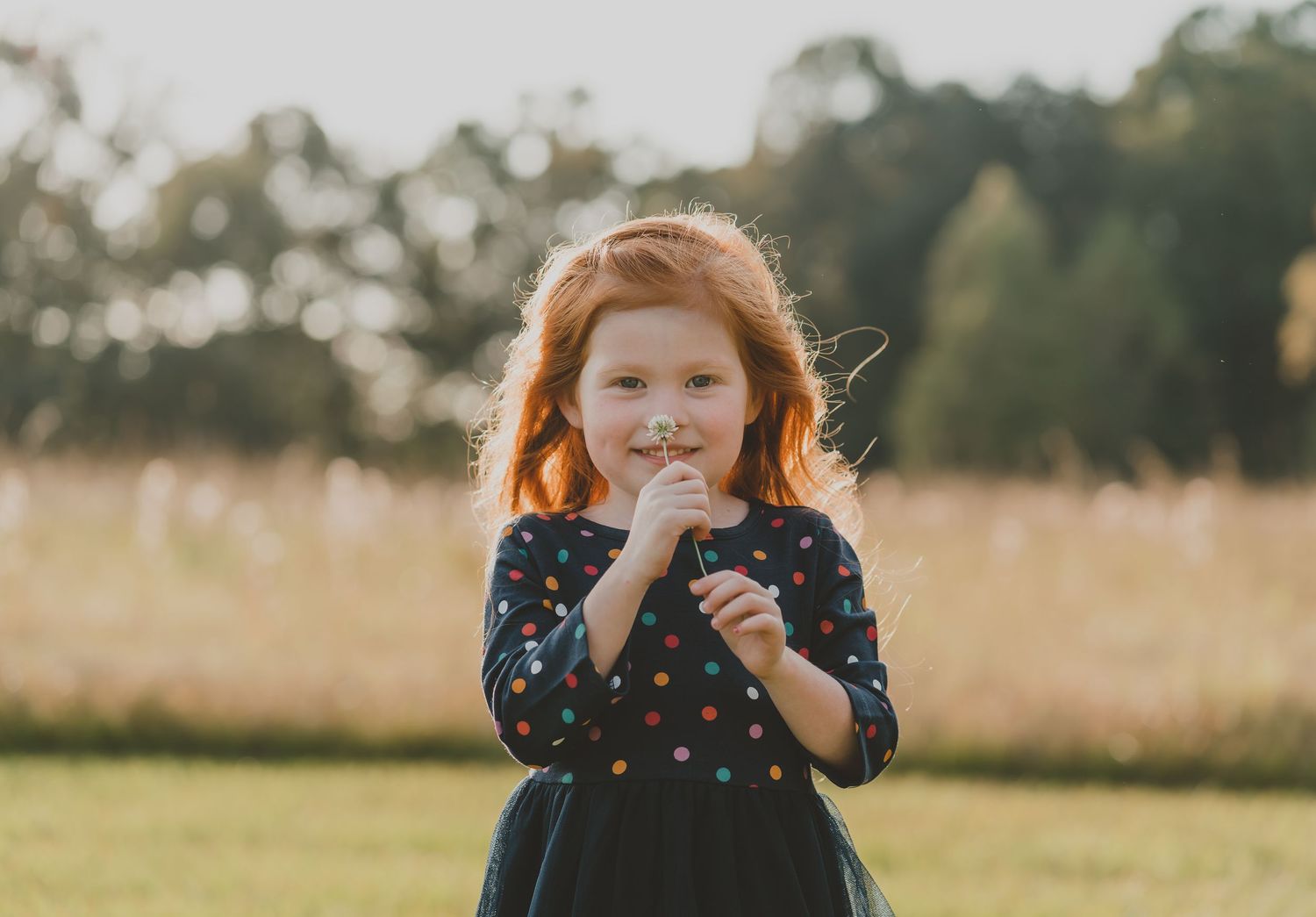 A child wearing a navy polka dot dress smiles playfully in a golden field at sunset.