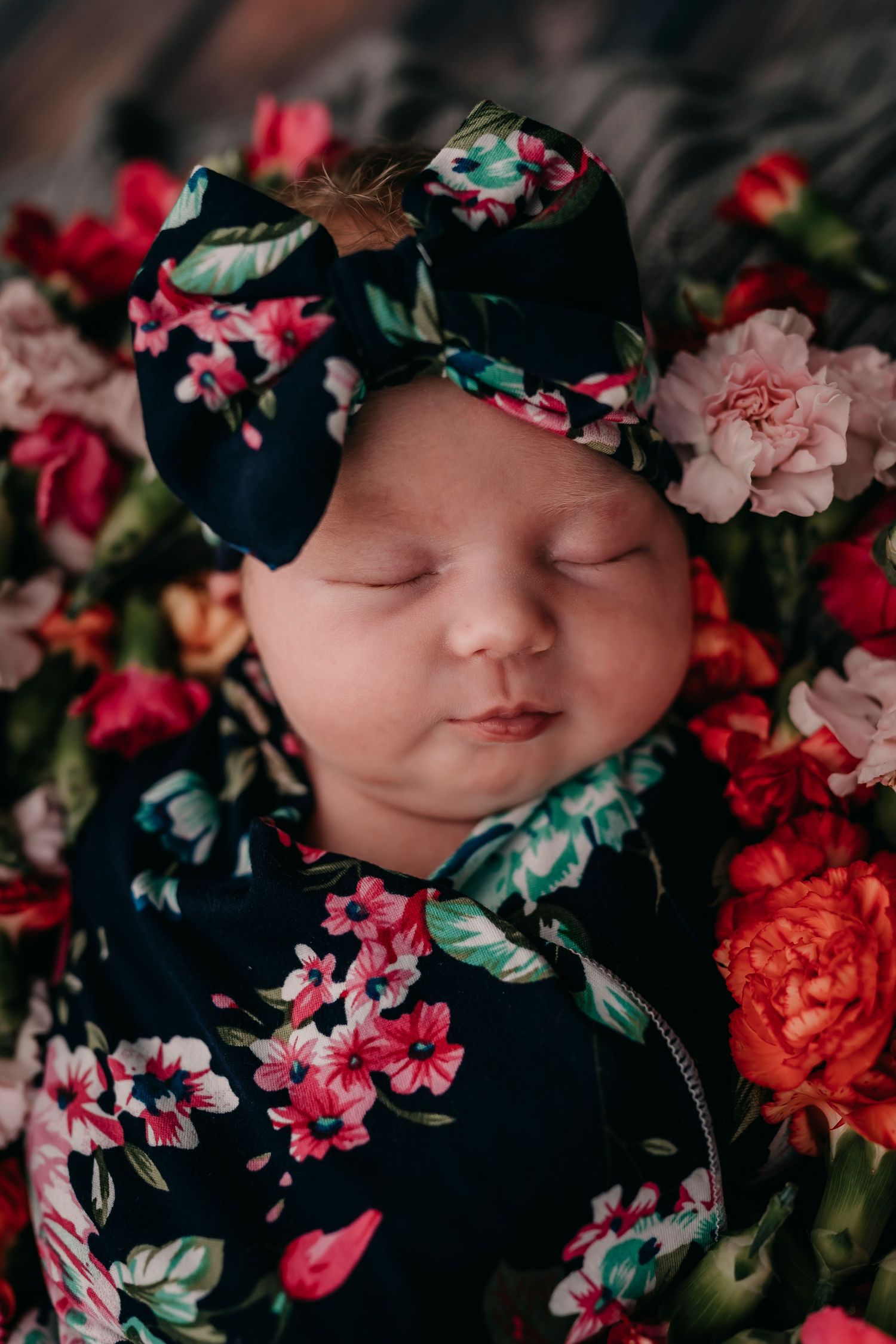 Close-up of sleeping baby in floral outfit and matching bow surrounded by pink roses.