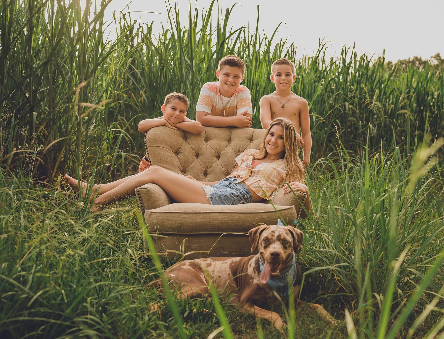 Group lounging on vintage couch placed in tall grass field with dog resting nearby.