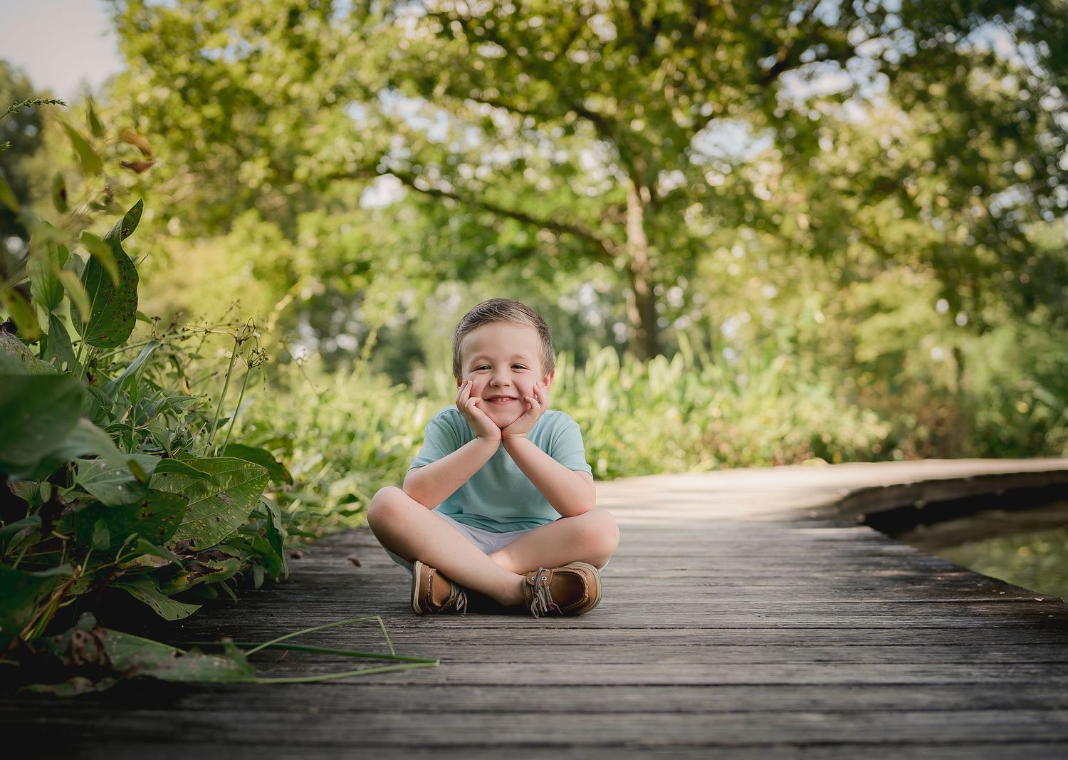Joyful outdoor portrait sitting cross-legged on a wooden pathway surrounded by greenery.