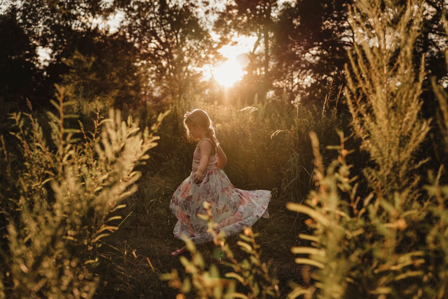Person in floral dress twirls among tall grass during sunset.