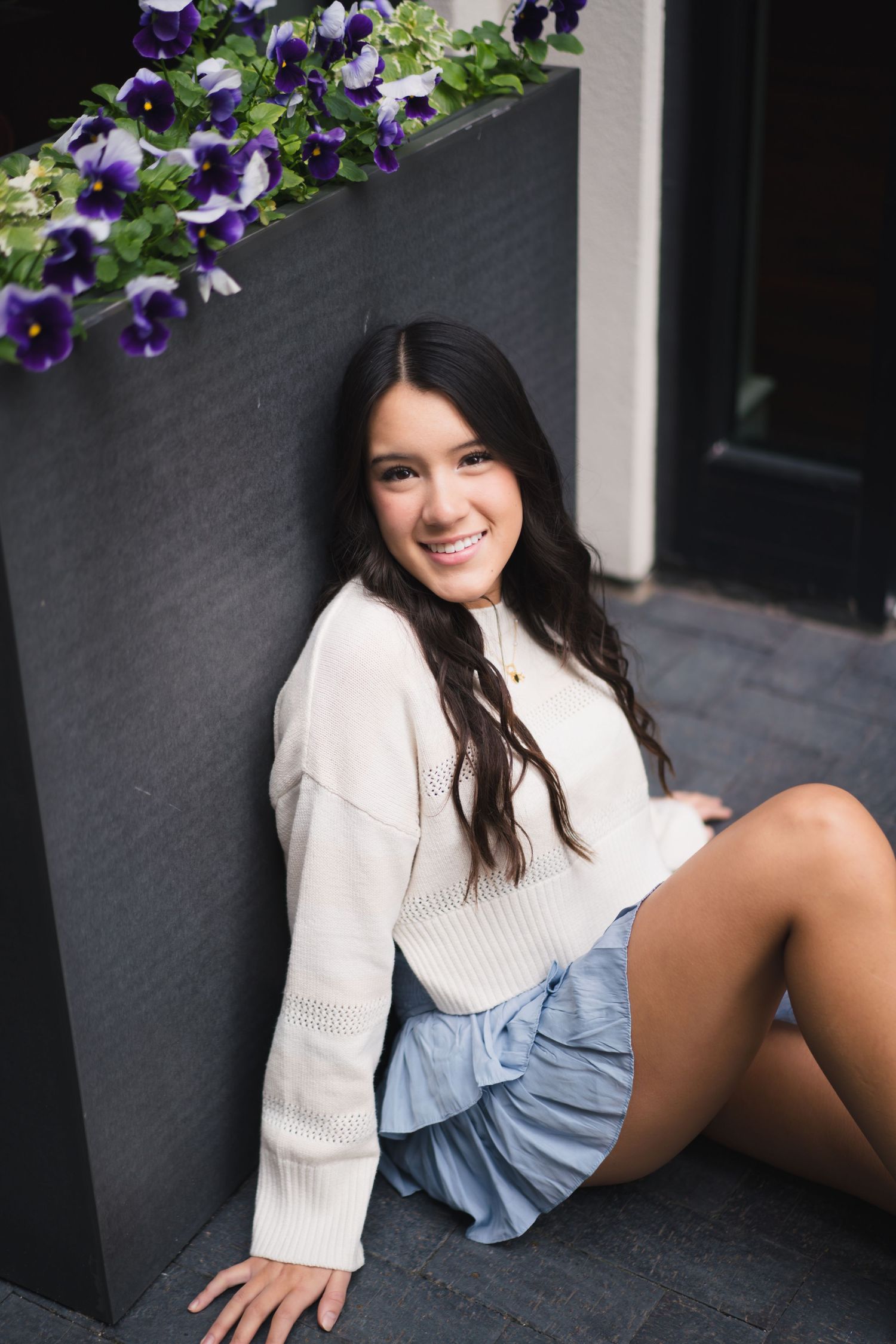 A woman in a white sweater and denim shorts lounges on a gray couch with purple flowers in the background.