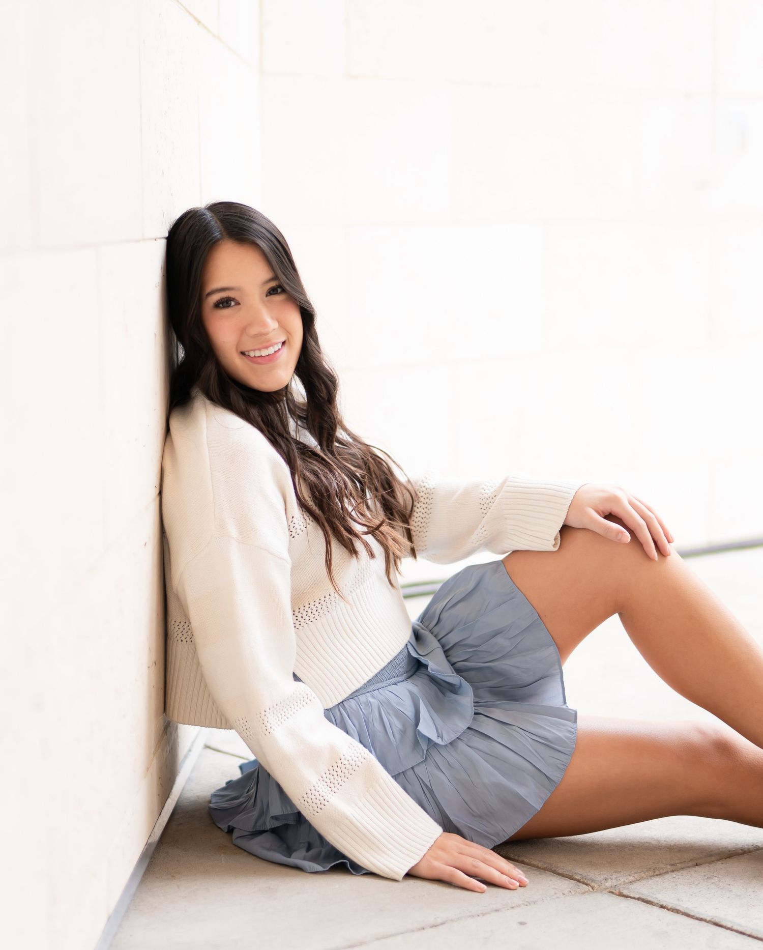 A person in a white blouse and gray skirt poses gracefully on a white couch.