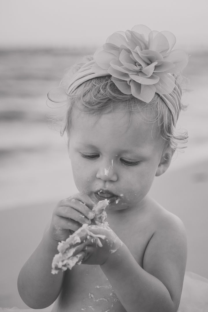 Black and white portrait of toddler with flower accessory enjoying messy cake.