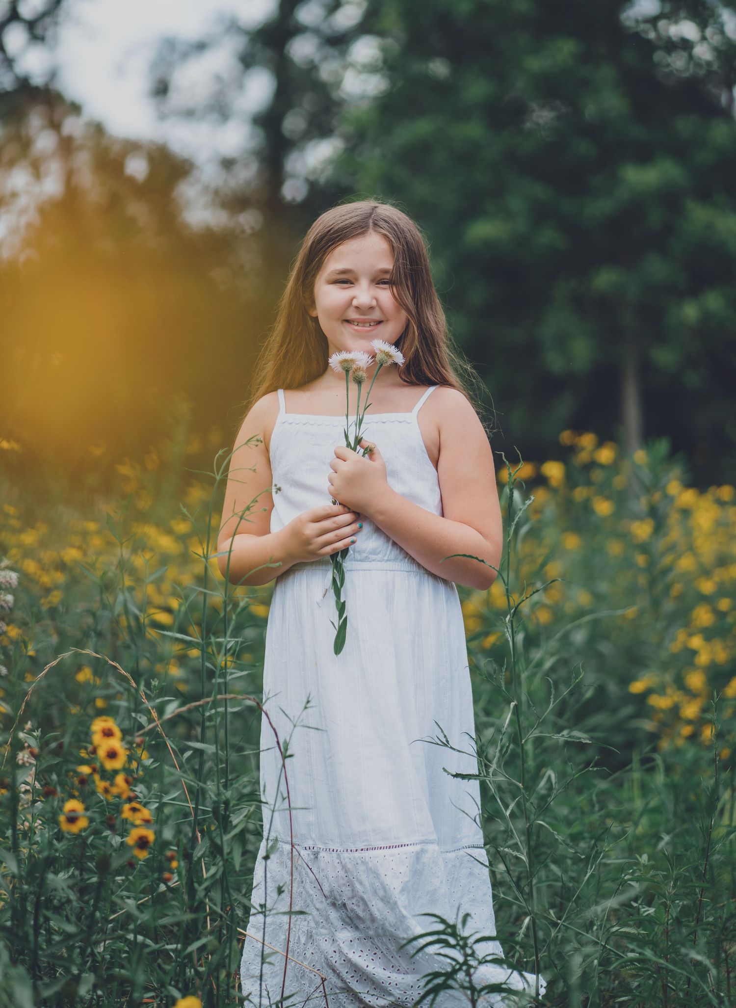 Young girl in white dress holds flowers while standing in field of yellow wildflowers.