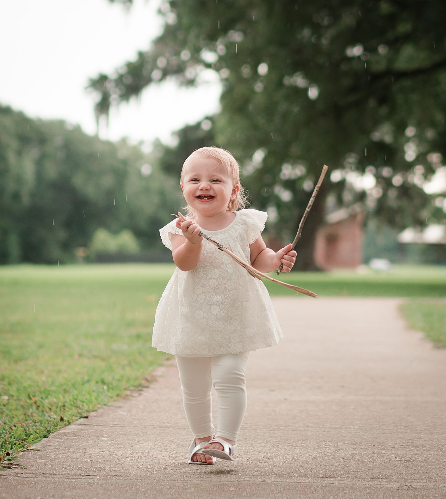 Small child in white dress walking on path holding stick with joyful expression.