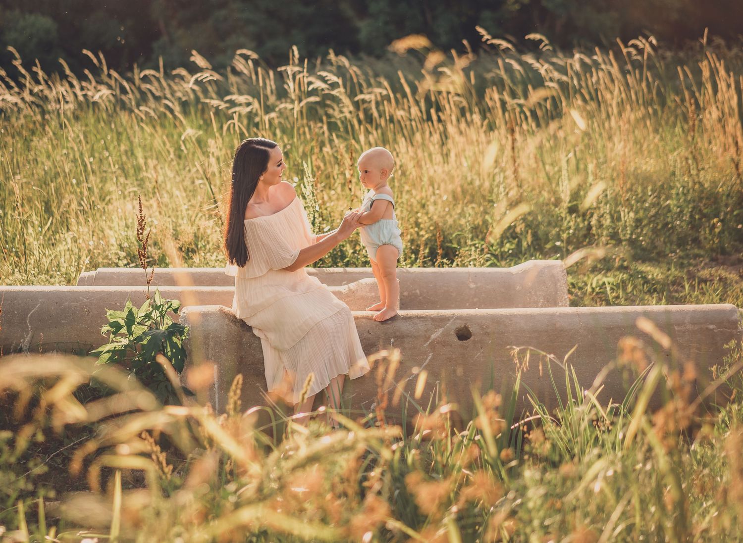 A mother and baby sit together on outdoor steps surrounded by tall golden grasses at sunset.