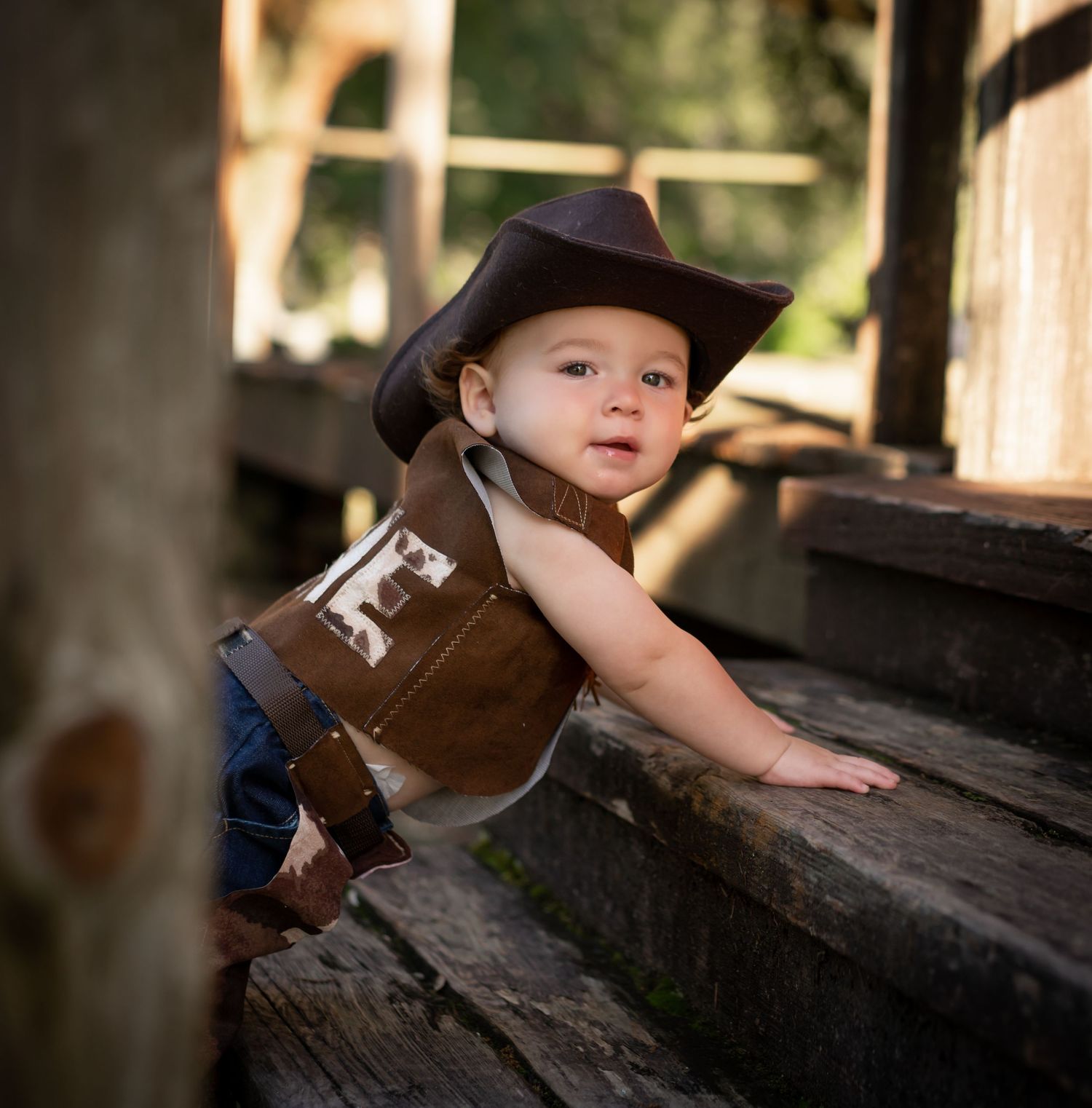 A baby wearing a cowboy hat and western vest crawls on wooden steps outdoors.