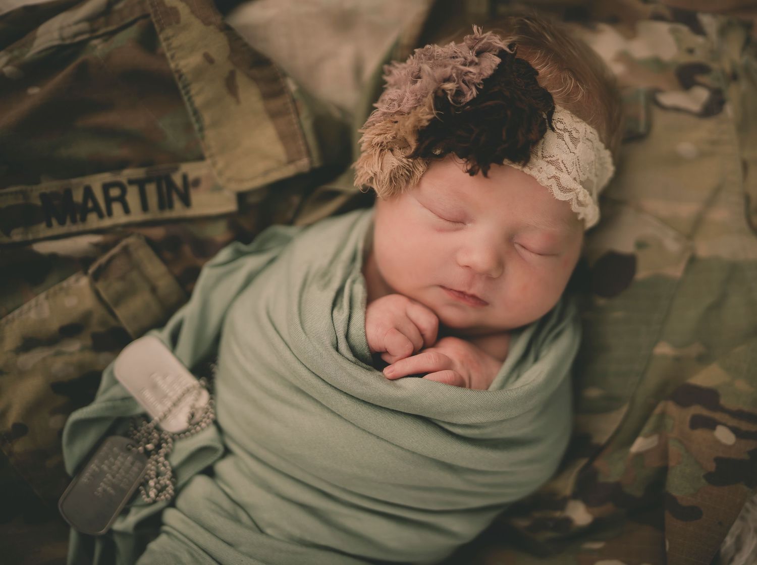 Sleeping infant wrapped in sage green swaddle on camouflage military uniform.