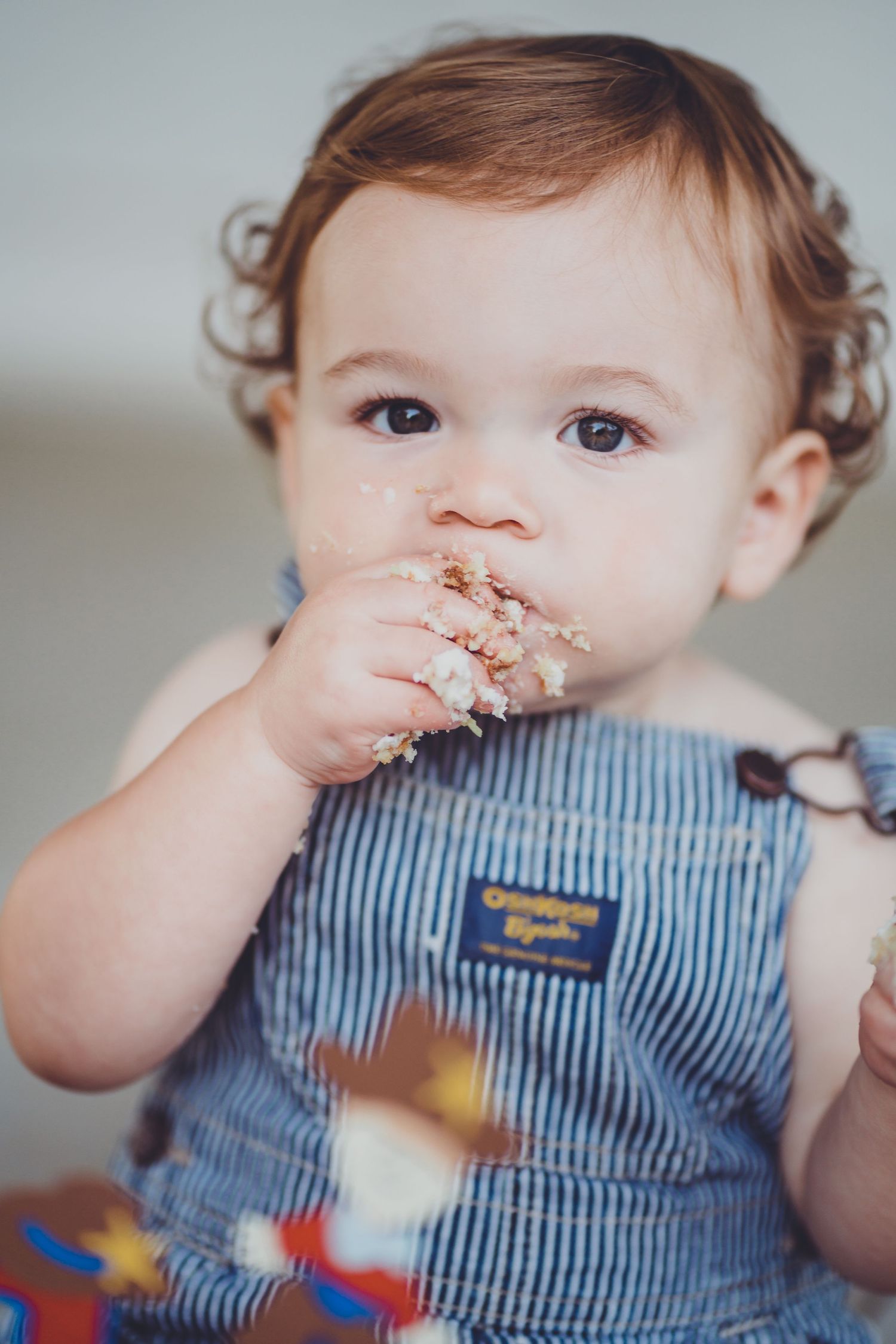 Close up of toddler in striped overalls eating cake with messy hands.