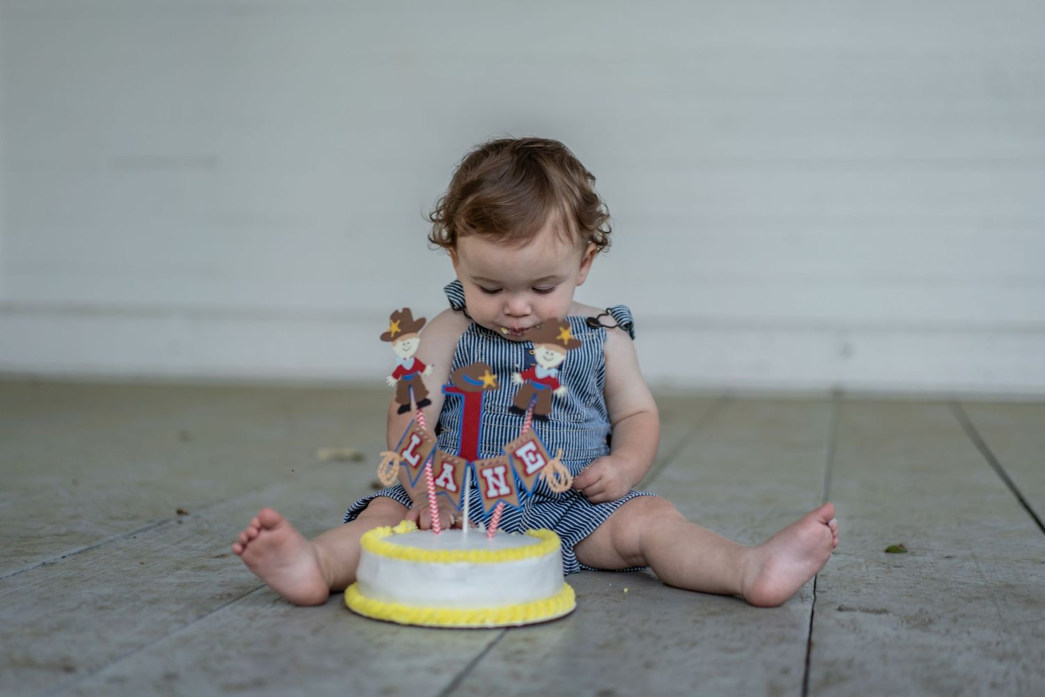 Small child in blue outfit sits on floor with birthday cake and decorative banner.
