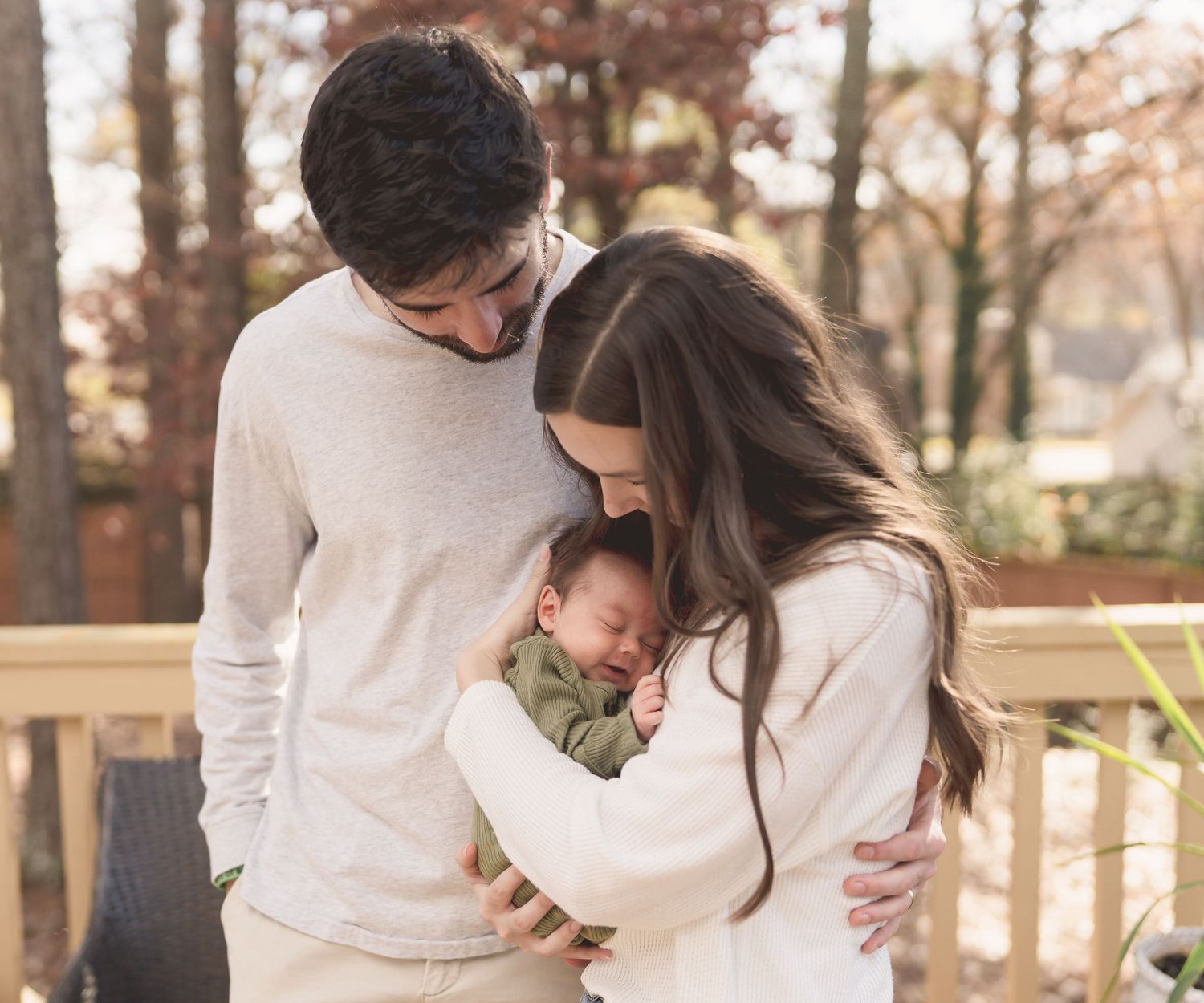 A tender family moment captured outdoors on a wooden deck during autumn with warm neutral tones and fall foliage.