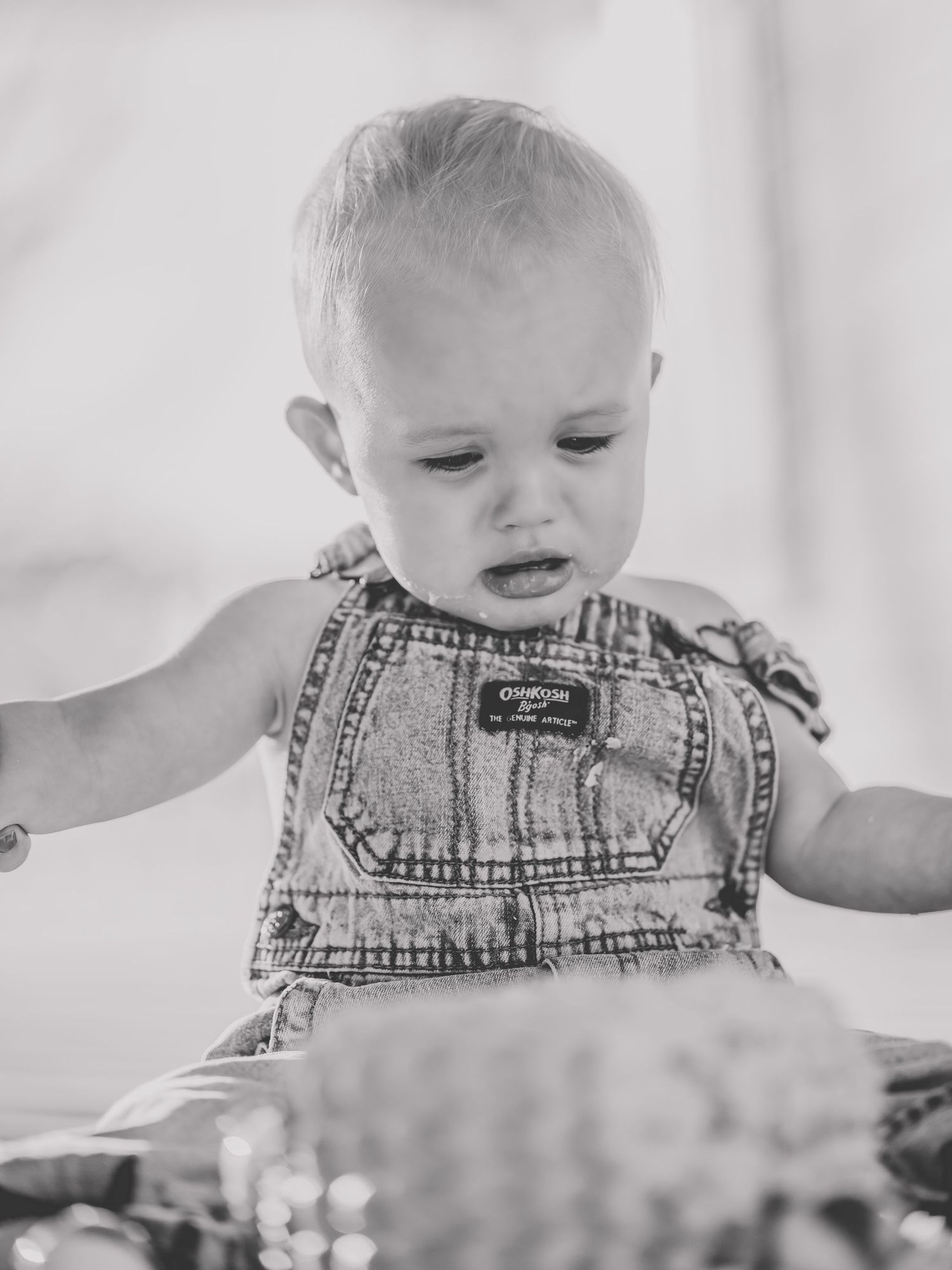 Black and white image of baby in denim overalls looking down at cake.