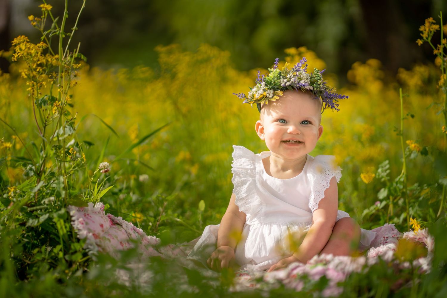 Joyful baby in white dress wearing floral crown sits among yellow wildflowers in garden setting.