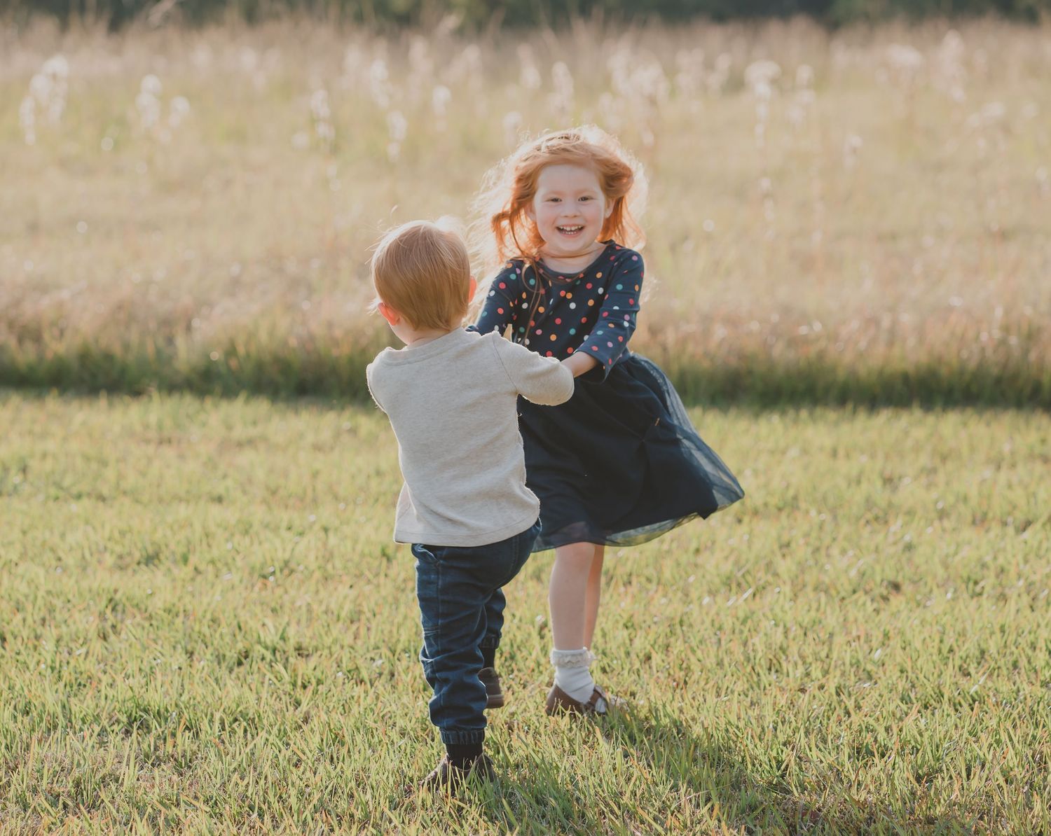 Two children playing and dancing together in sunlit grassy field.