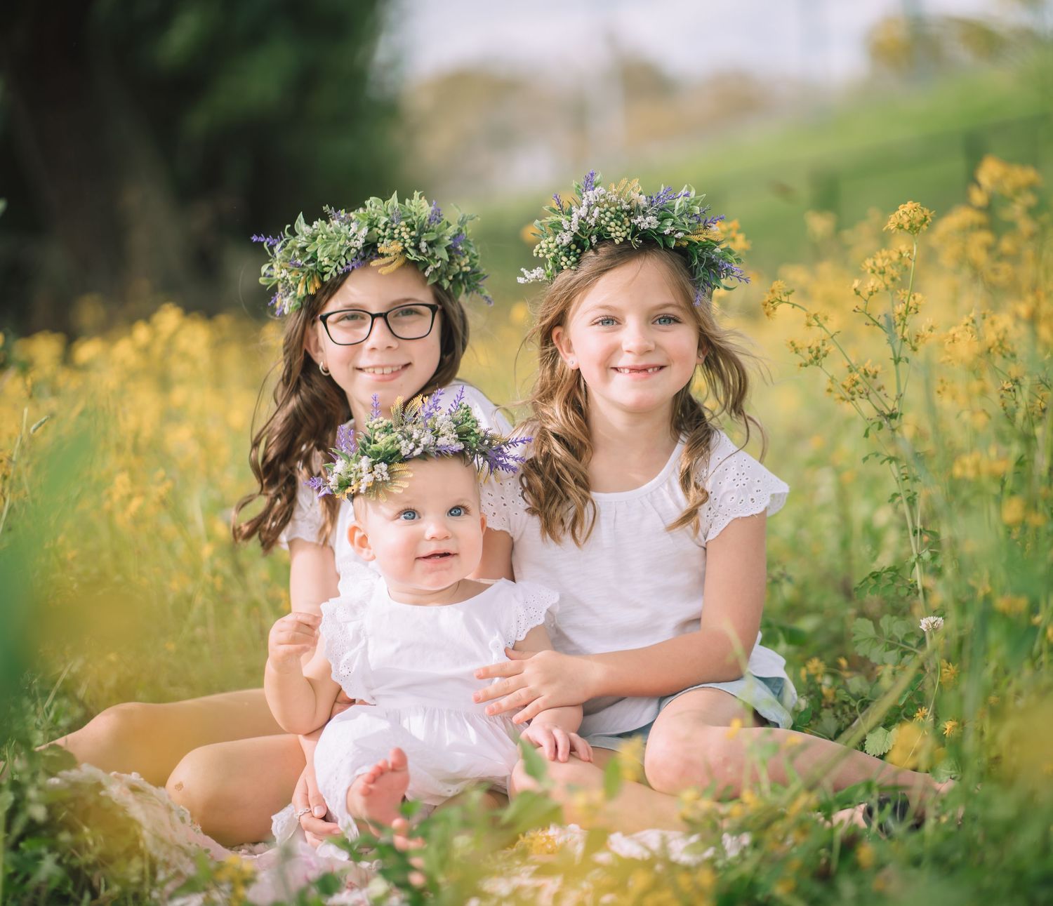 Three siblings wearing matching floral crowns pose together in a field of yellow wildflowers.