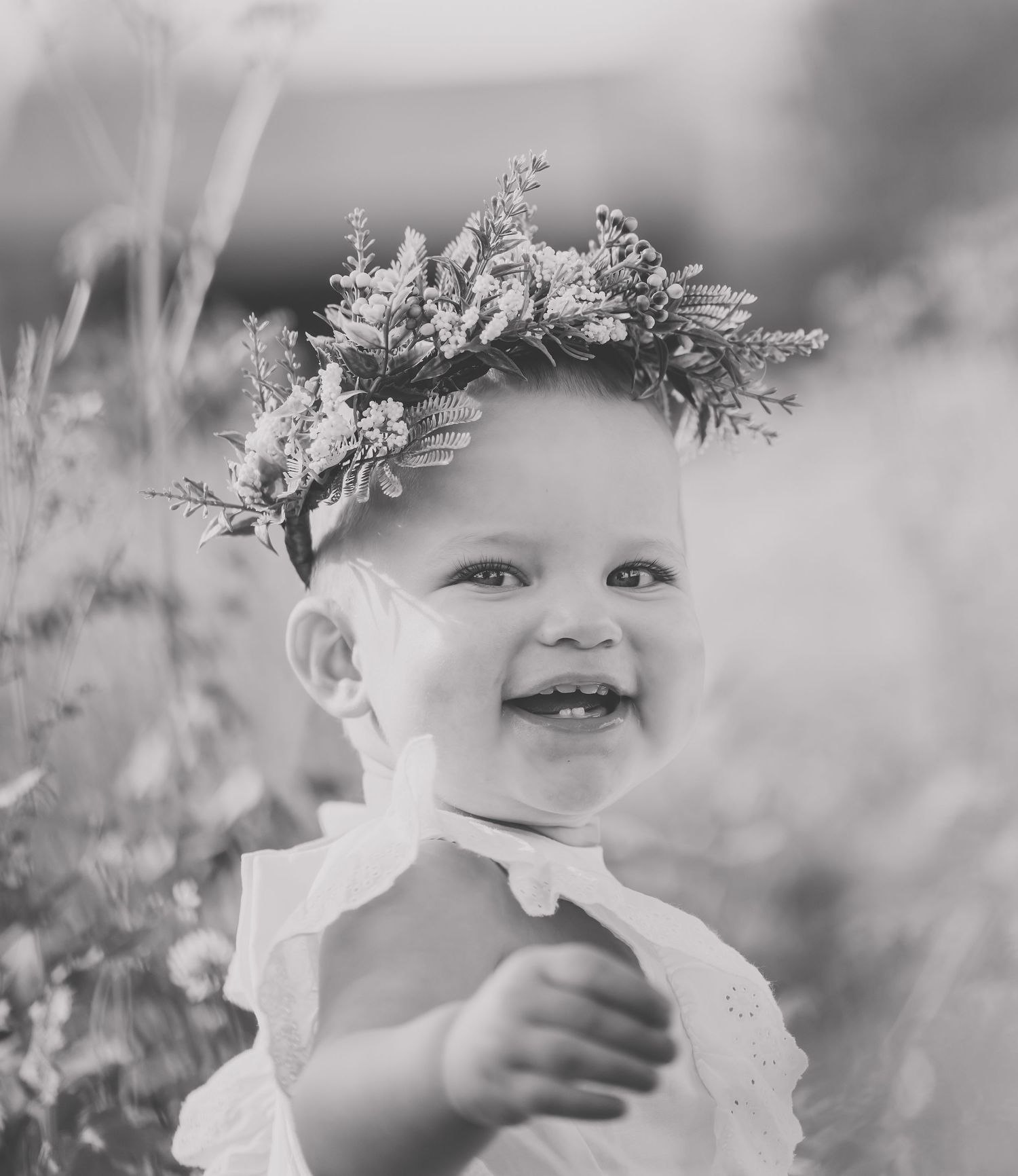 Black and white portrait of a smiling baby wearing a delicate floral crown and white dress.