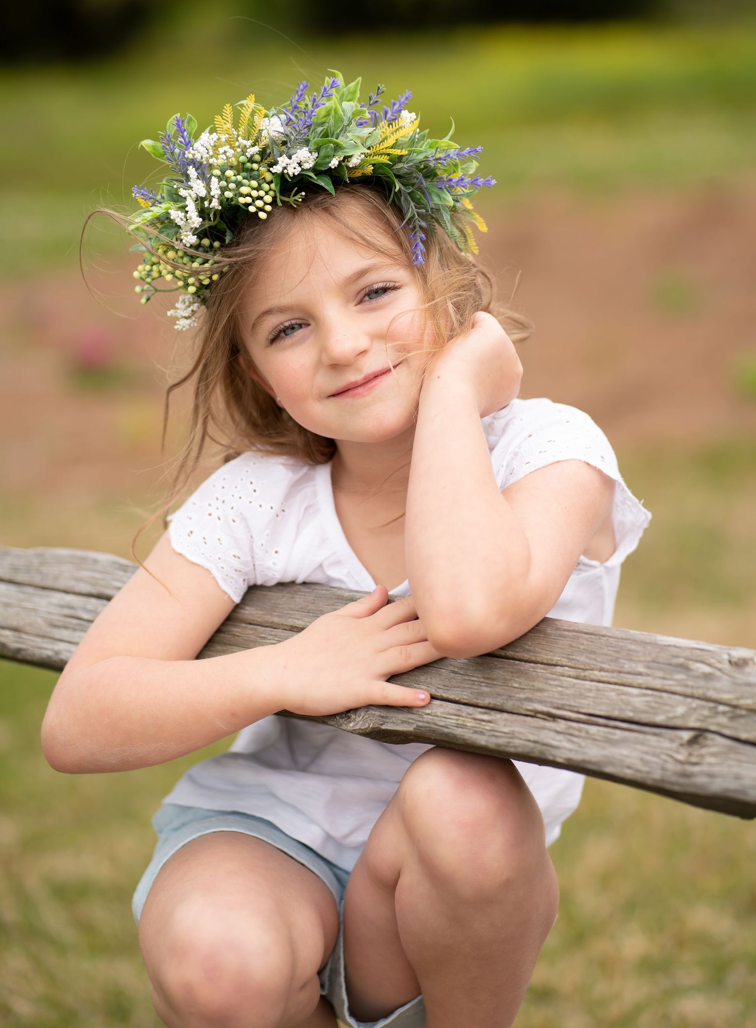 Whimsical outdoor portrait of a child wearing a floral crown while leaning on a wooden fence in nature.