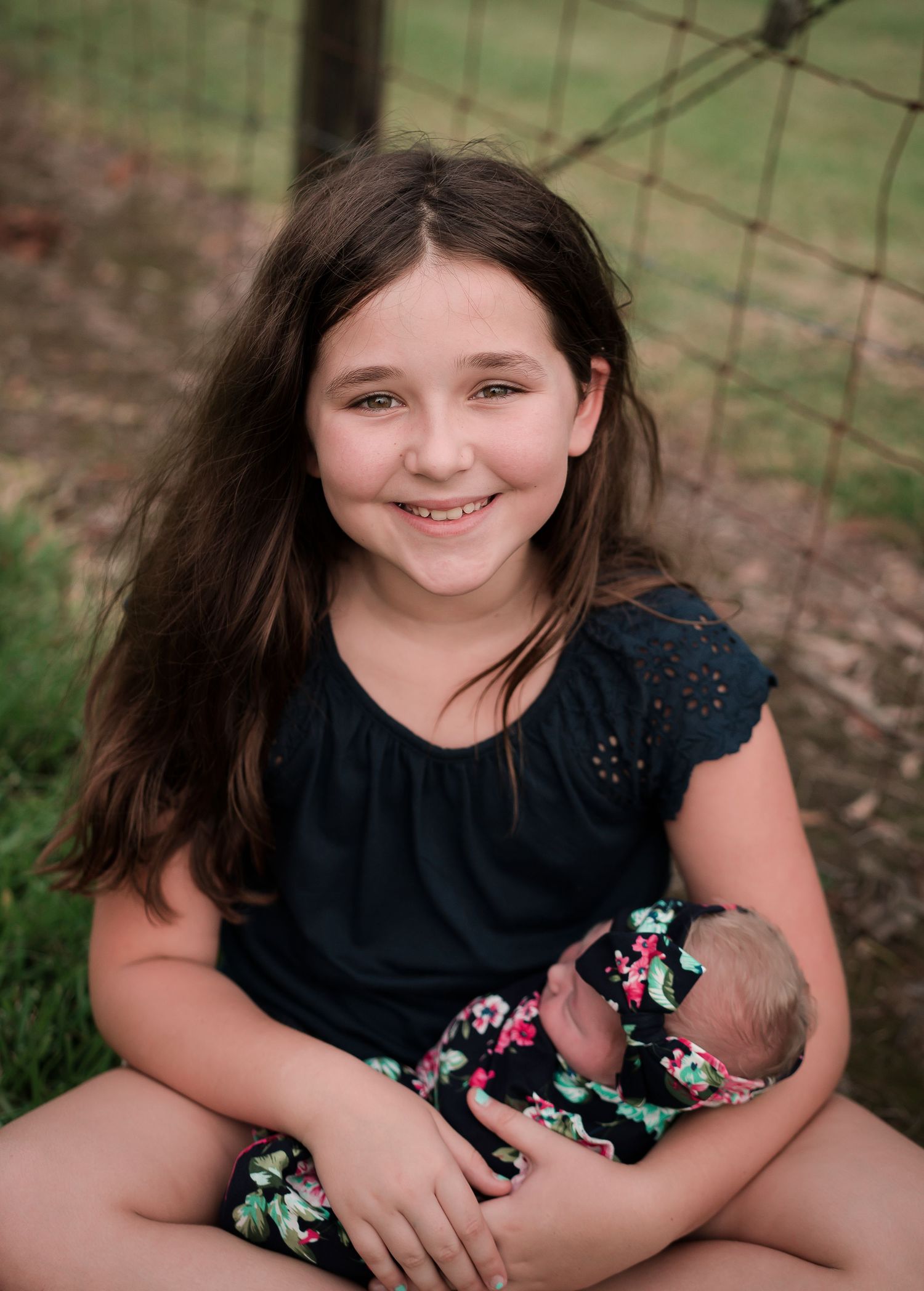 Young person in navy blue top holding small baby in floral outfit near rustic fence.
