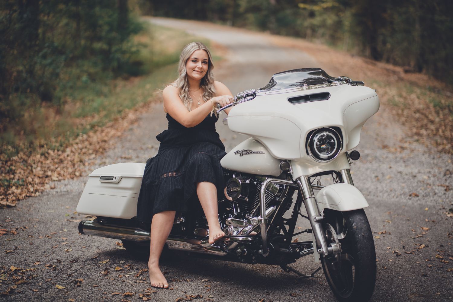 A motorcycle rider poses with a custom white cafe racer motorcycle on a leaf-covered forest road.