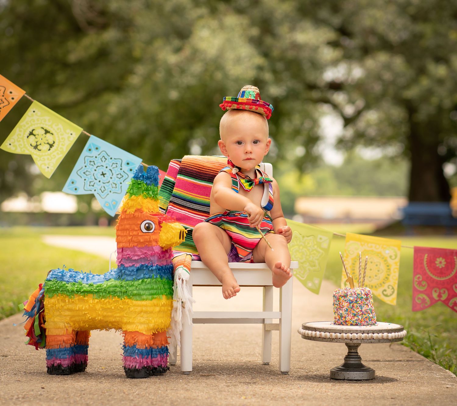 Baby sits on white chair with colorful pinata and birthday cake at outdoor party.