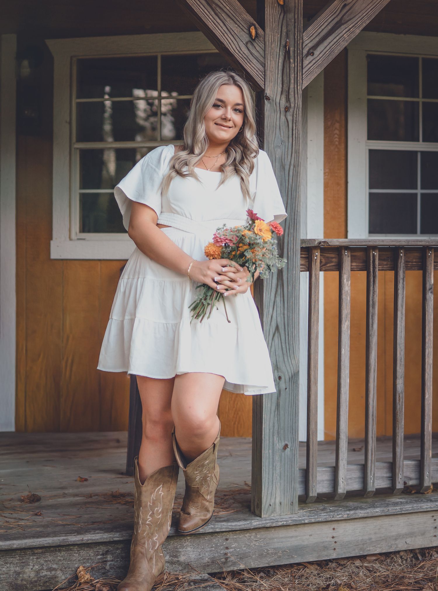 Someone in a white flowy dress and brown cowboy boots holds a colorful bouquet while standing on a rustic wooden porch.