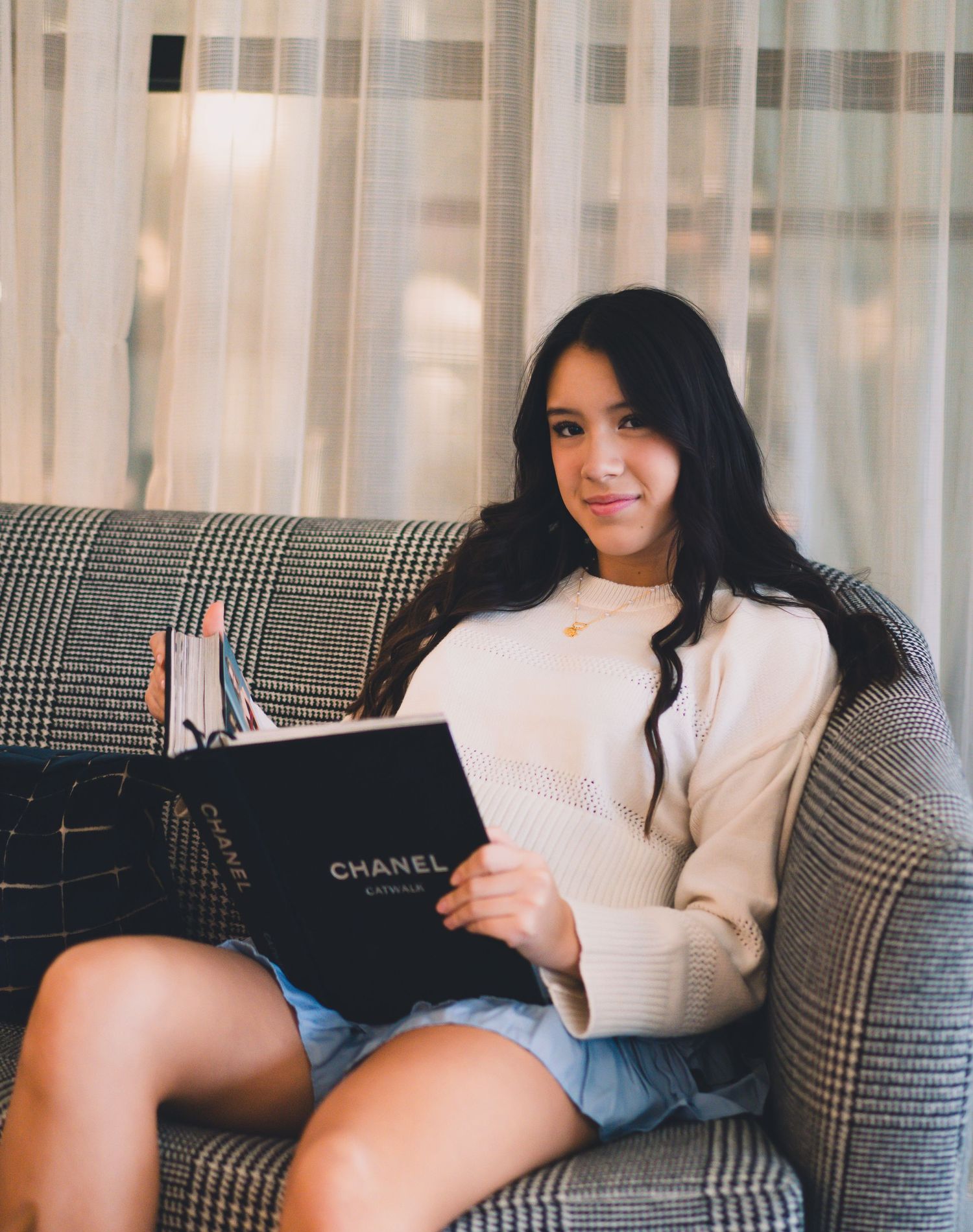 Someone relaxes on a plush gray couch while reading a Chanel book, wearing a cozy white sweater and denim shorts.