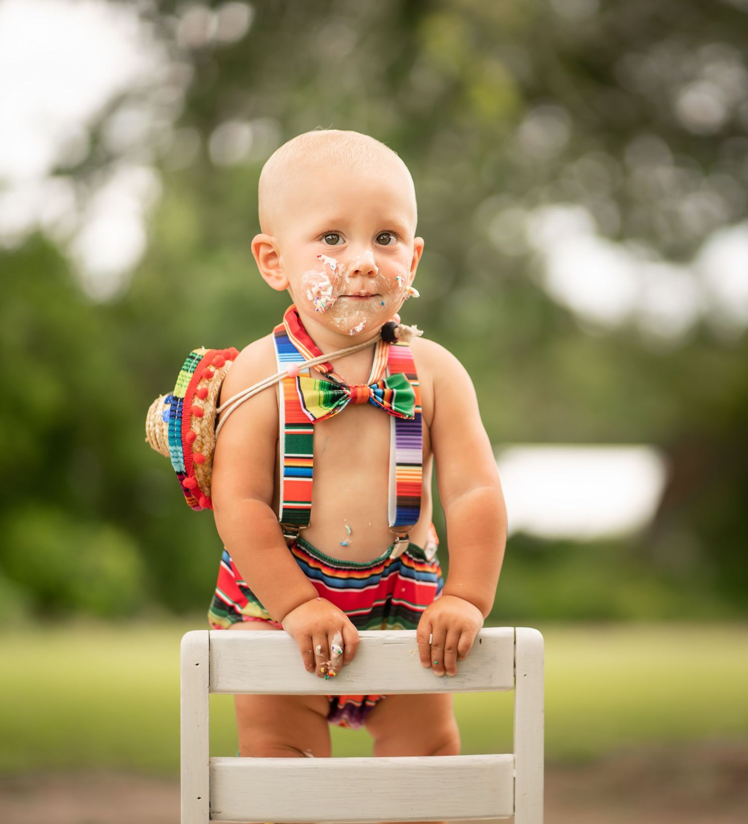 Baby in striped suspender outfit stands on white chair with frosting on face.