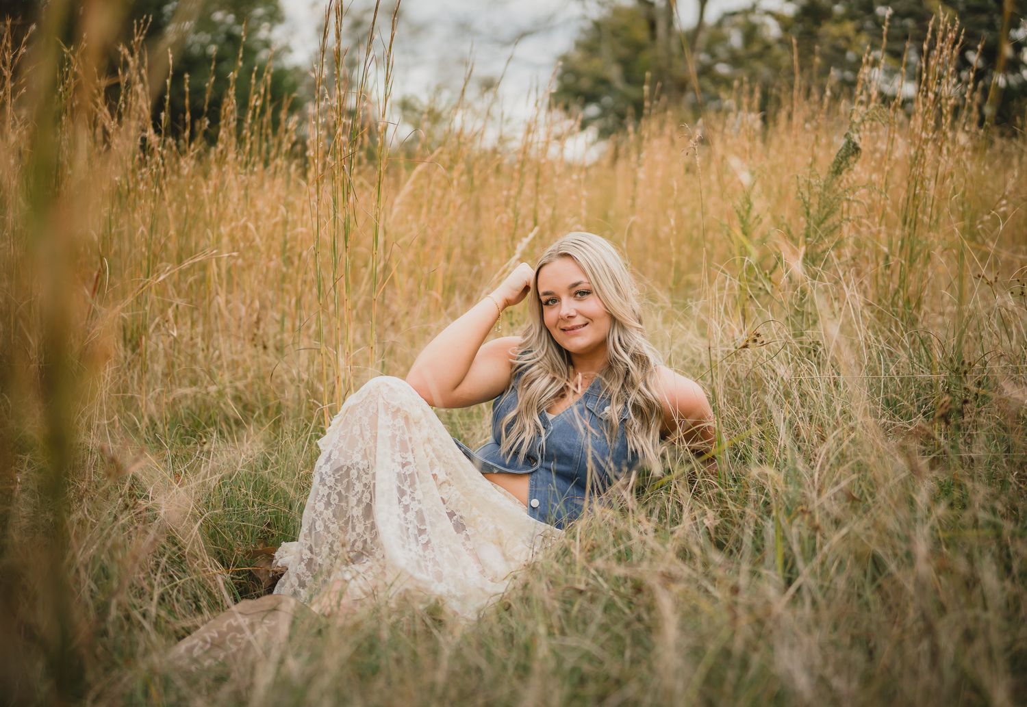 Woman relaxes in tall grass meadow holding fresh picked flowers during golden hour photoshoot.