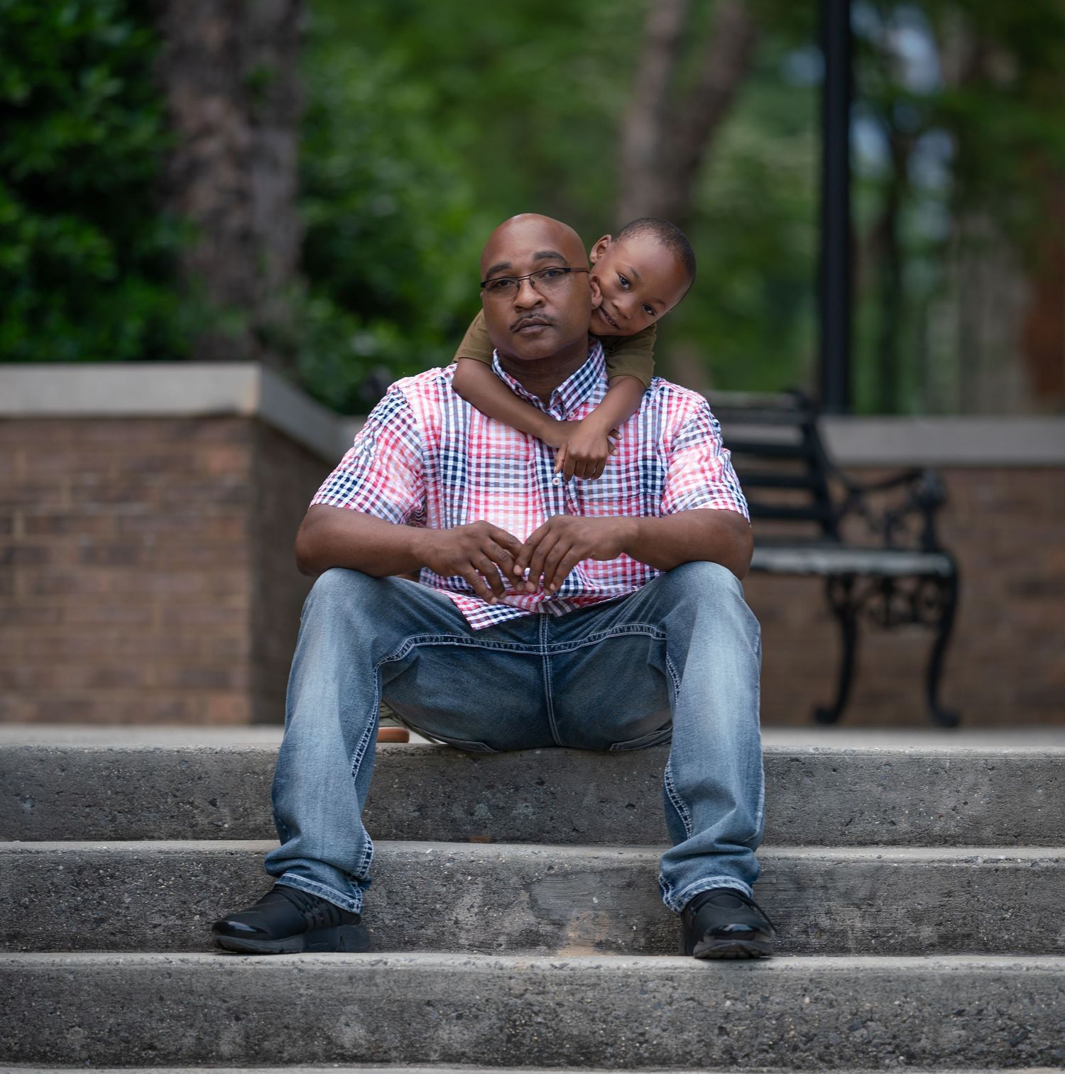 Two people share a tender moment sitting on concrete steps outdoors with greenery and a brick wall in the background.