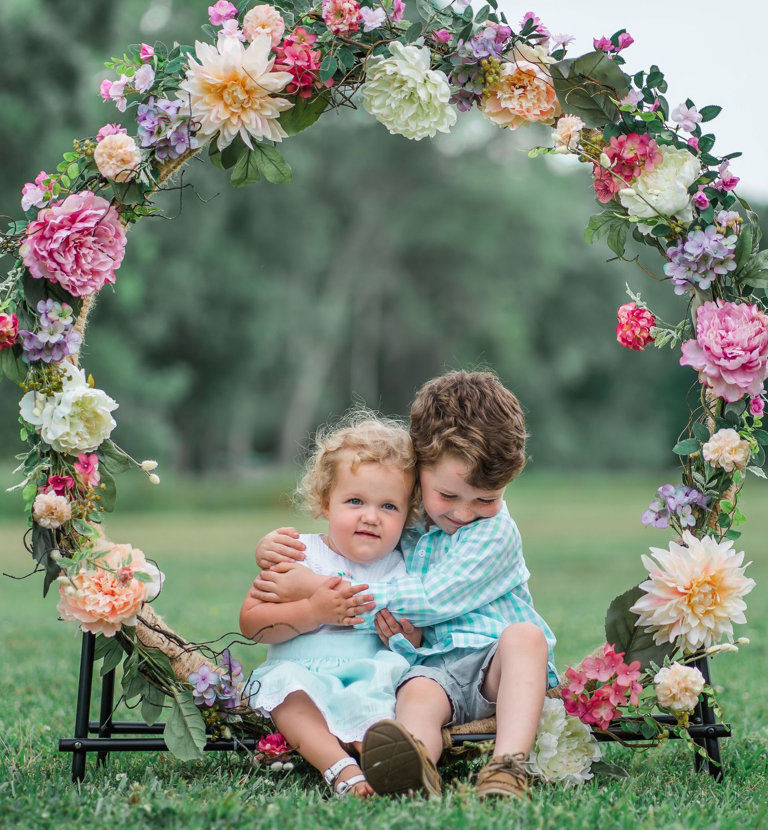 Children embrace under gorgeous floral arch during outdoor spring photo session.