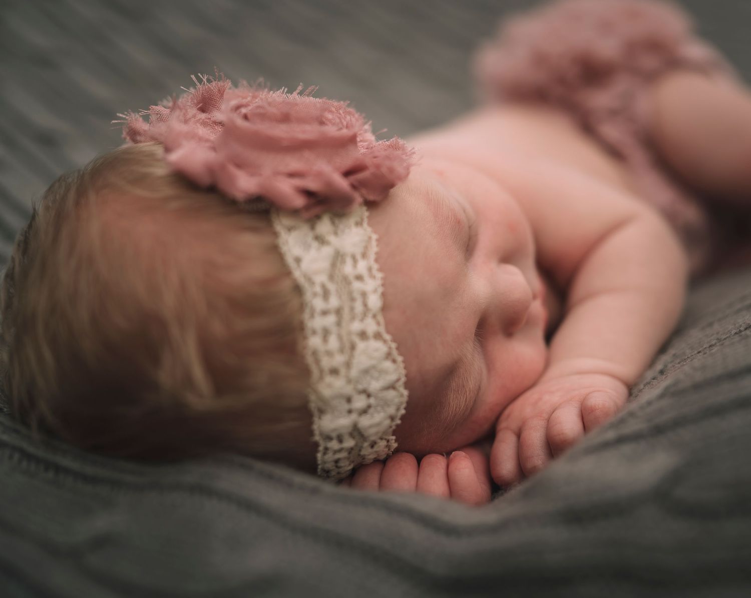 Sleeping baby wearing pink flower headband on gray blanket during newborn photoshoot.
