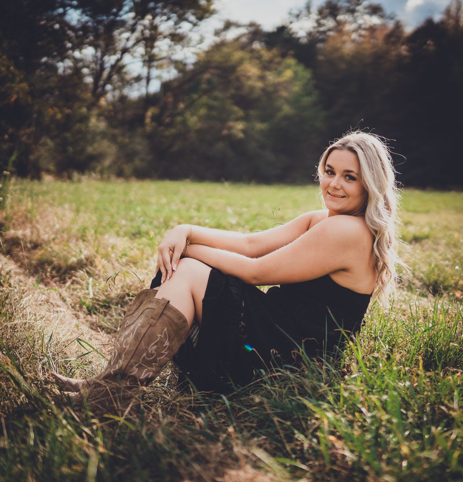 Person in black dress lying gracefully in a sunlit meadow with tall grass at golden hour.