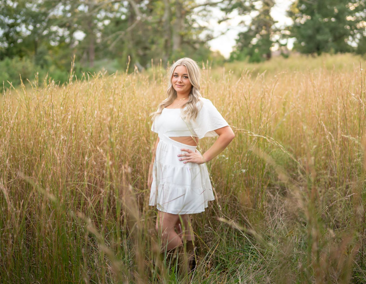 A figure stands in a golden field of tall grass during sunset wearing a flowing white top and casual attire.