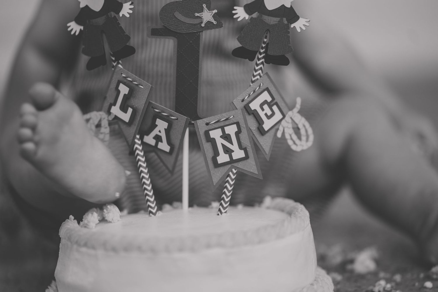 Black and white detail shot of birthday banner and cake with small feet in background.