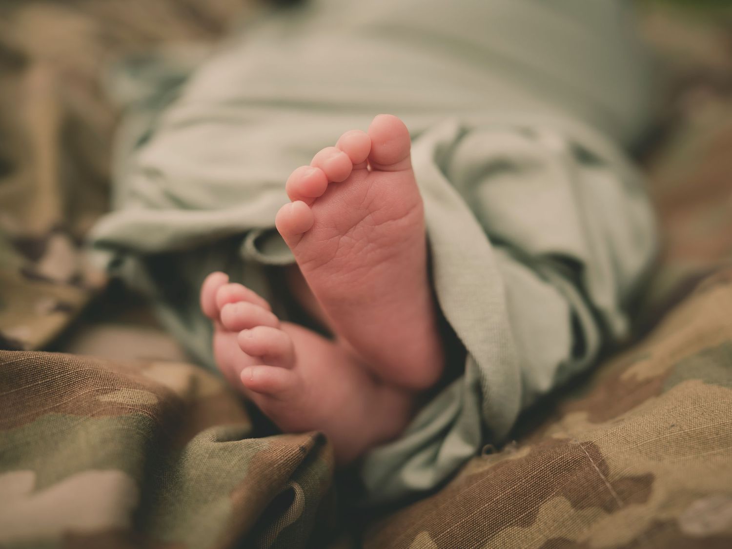 Tiny baby feet peek out from sage green swaddle against camouflage military uniform background.