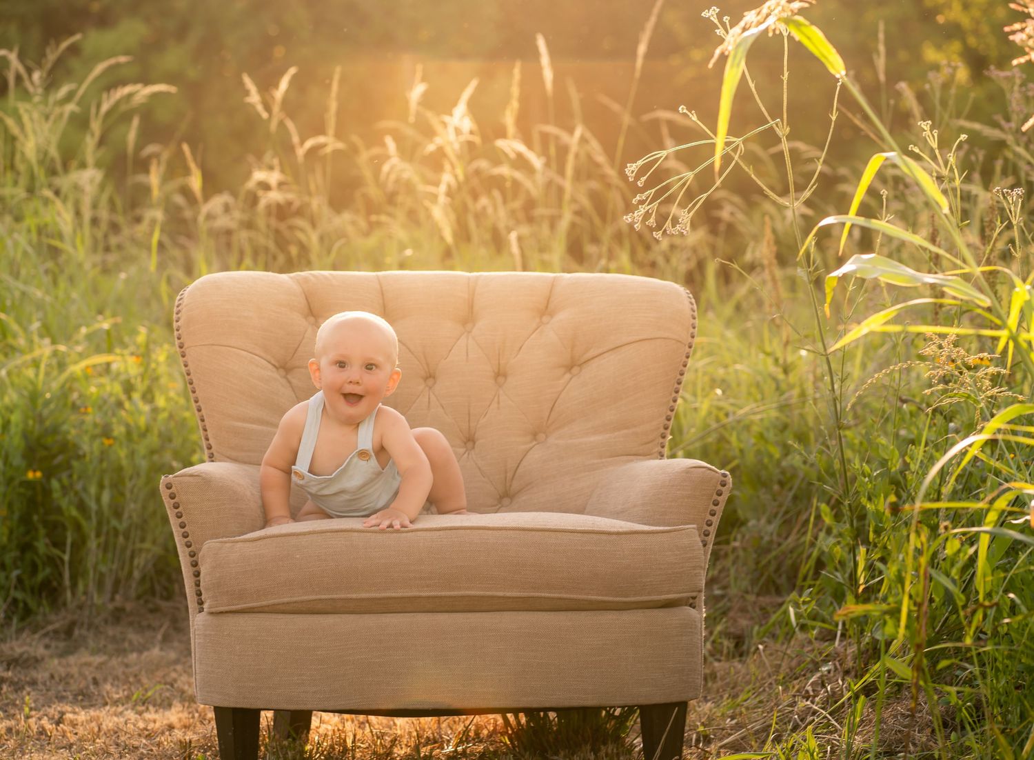 A baby sits and smiles in a beige tufted armchair outdoors amid tall grass.