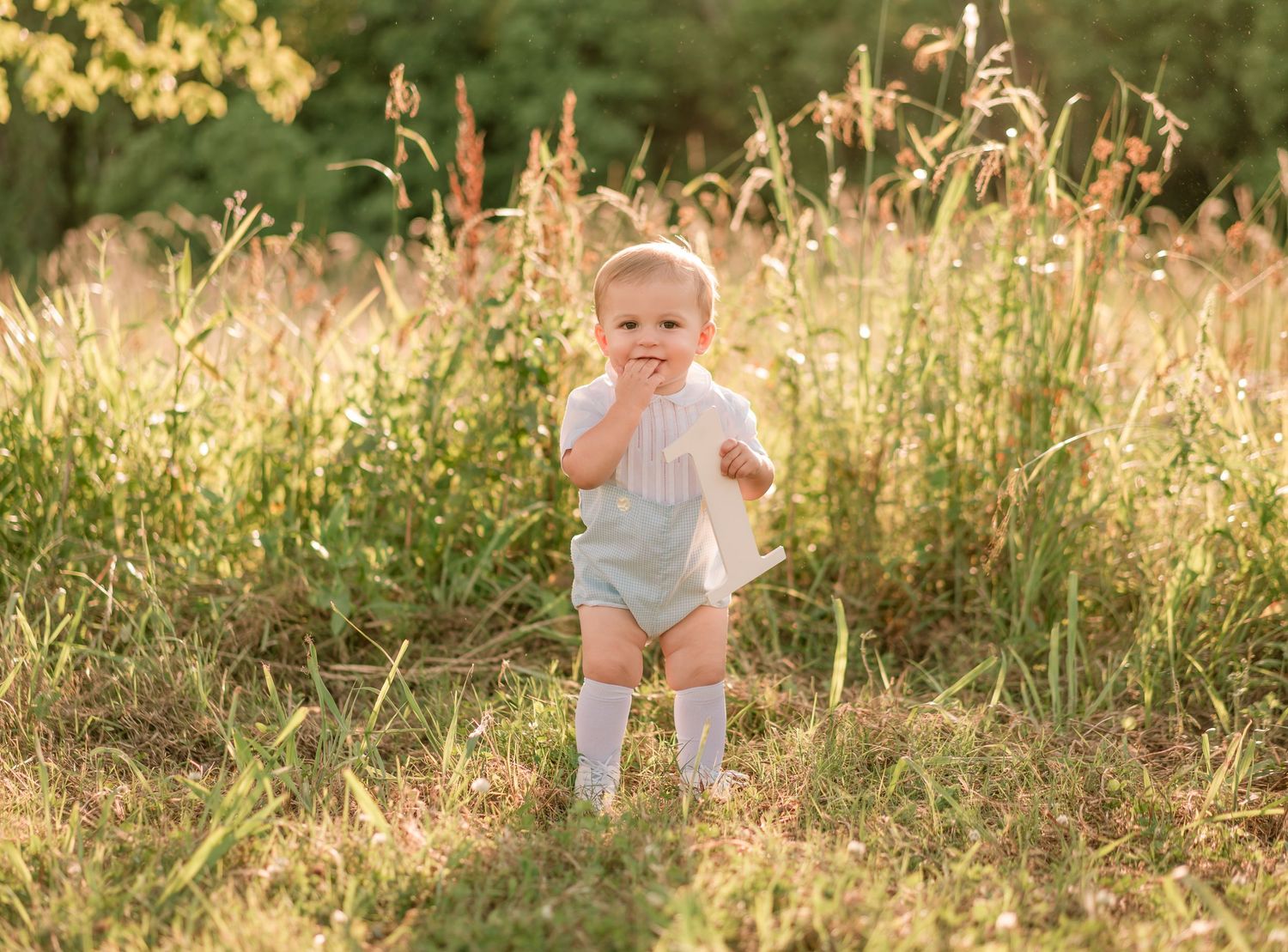 A baby in a light colored romper stands among golden grass during sunset.